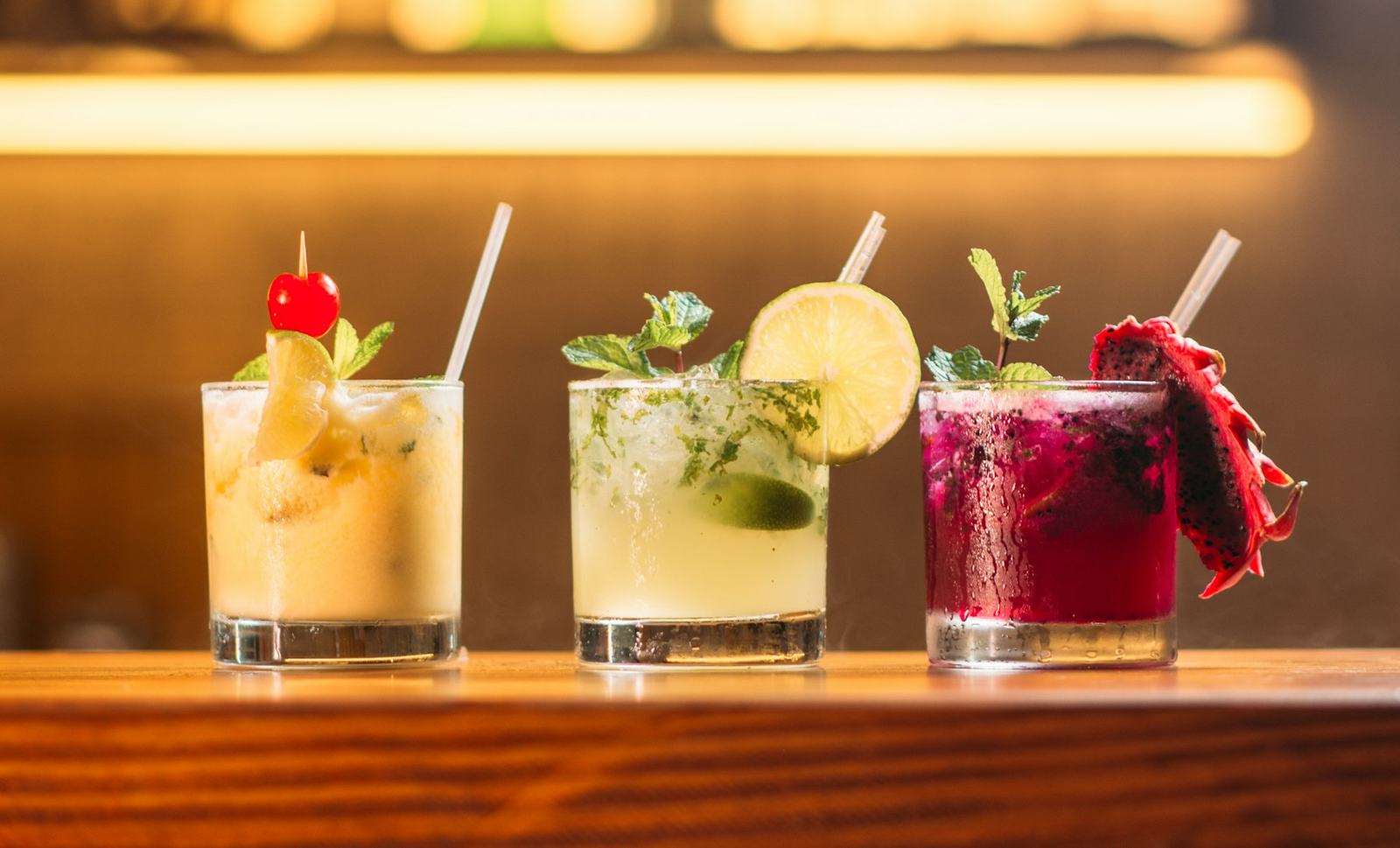 Three finished cocktails lined up on a bar counter under warm strip lighting